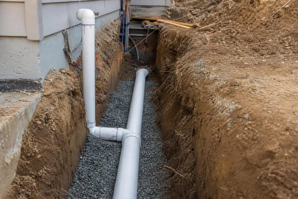 White PVC pipes installed in a deep dirt trench with gravel bedding alongside a residential building foundation.